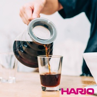 一個玻璃咖啡壺正在將深棕色咖啡倒入一個透明玻璃杯中，咖啡壺帶有銀色金屬濾網和透明瓶蓋。