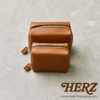 兩款大小不同的職人手作皮革箱型收納包，皆為棕色，搭配金色拉鍊與同色皮革拉環，擺放在灰色紋理背景上，下方有HERZ品牌標誌與HAND MADE IN JAPAN Since1973字樣