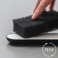 一隻手正拿著黑色網狀海綿，海綿上方有細小的水珠，下方是一個帶有白色內襯的黑色長方形皂盤，背景為淺灰色。