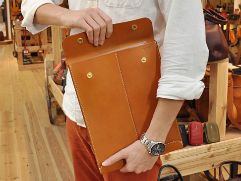 職人手作皮革信封式雙釦手拿袋，採用鞣製皮革，顏色為大地棕色，配有金色釦子和白色縫線，設計簡約大方。