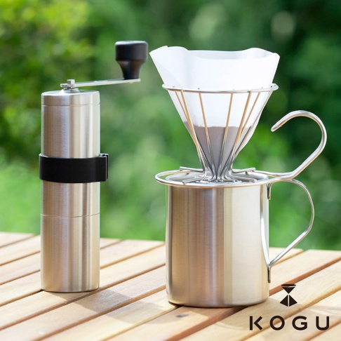 日製18-8不鏽鋼手搖咖啡磨豆器，搭配同材質的濾杯架及帶有手柄的咖啡杯，磨豆器上有黑色防滑帶，背景為模糊的綠色植物。