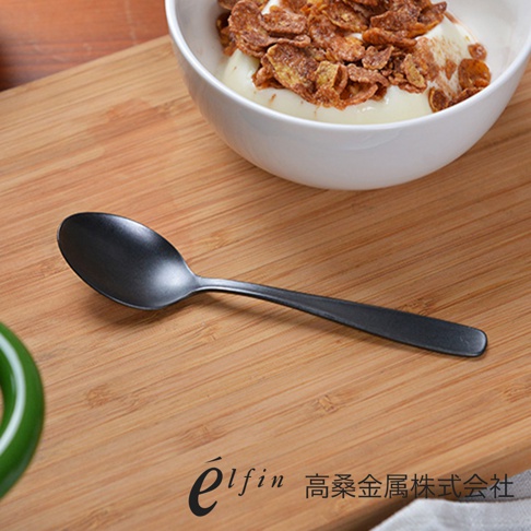 日本製熔岩黑石紋理水果甜點匙，啞光黑色金屬材質，勺頭呈橢圓形，勺柄細長，放置於木質桌面，背景為裝有麥片和優格的白色碗。