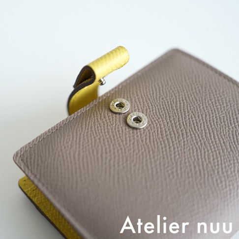 Atelier nuu 品牌標誌出現在灰色皮革短夾的右下角，短夾上有兩個銀色金屬按扣。