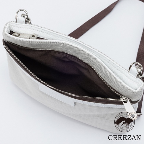 一款 CREEZAN 品牌的白色義大利帆布拼接皮革斜背包，採用高角度俯視視角，展示了打開後的棕色內襯與銀色金屬拉鍊細節，包款邊緣以白色皮革包邊，並配有棕色背帶，右下角印有品牌標誌與名稱。