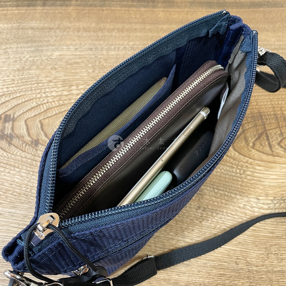 藍色刺子織斜背包，內部有手機、錢包及綠色小物，背包開口拉鍊與肩帶為黑色。