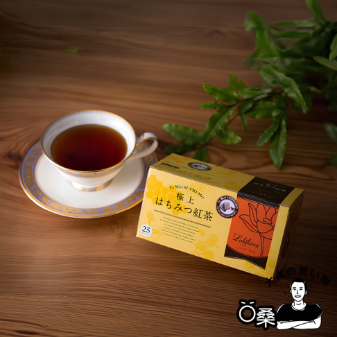 Lakshimi 極上蜂蜜紅茶盒裝，搭配一杯熱氣騰騰的紅茶，置於木質桌面，旁有綠葉裝飾