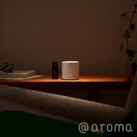 淺灰色圓柱形隨身精油香氛機，頂部有圓形出霧口，側面有一盞小黃燈。旁邊擺放著一瓶深棕色精油瓶，瓶身標有「S 06」字樣。上方有一疊書籍和一副眼鏡。