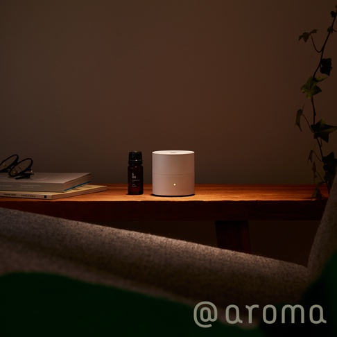 淺灰色圓柱形隨身精油香氛機，頂部有圓形出霧口，側面有一盞小黃燈。旁邊擺放著一瓶深棕色精油瓶，瓶身標有「S 06」字樣。上方有一疊書籍和一副眼鏡。