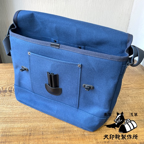藍色純棉帆布雙扣郵差包，配有黑色皮革裝飾和銀色金屬扣，包身正面有兩個筆插，肩帶可調節。