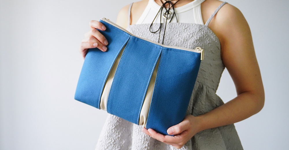 一位身穿灰色細肩帶洋裝的女性，雙手持著一個京都KOSHO品牌的ougi帆布化妝收納包。該收納包為長方形設計，外觀由藍色帆布與米白色布料交錯拼接而成，呈現獨特的扇形摺疊視覺感，配有金屬拉鍊，整體質感簡約優雅。