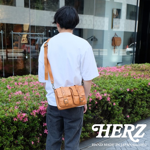 一位男性背著日本HERZ職人手作淺棕色皮革肩背包，包款設有兩個前置口袋，搭配白色短袖上衣與深色牛仔褲，右下角印有HERZ品牌商標及標語，背景為花圃與店面。