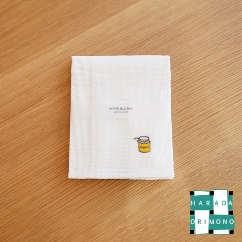 白色紗織紋擦拭巾，印有日文「かや生地ふきん」和英文「KAYA KIJI FUKIN」字樣的紙帶，右下角繡有黃色蜂蜜罐圖案，背景為木紋桌面。