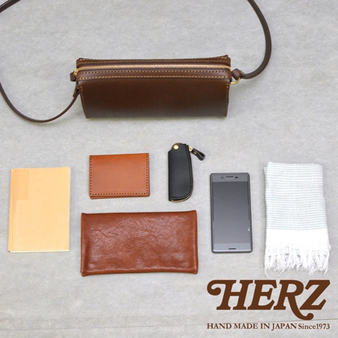 俯視視角下展示一個深棕色日本HERZ手作皮革單肩包，包袋上方放置了日常攜帶物品，包括一本黃色筆記本、棕色皮革名片夾、黑色皮革鑰匙包、棕色長皮夾、一台黑色智慧型手機以及一條淺灰色條紋毛巾，右下方印有HERZ品牌商標與手作標語。