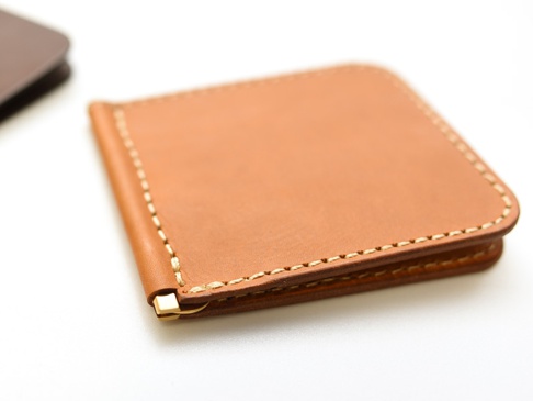 職人手作皮革橫式對折鈔票短夾，採用淺棕色皮革，搭配米白色縫線，夾角處設有金色金屬夾，簡潔實用。