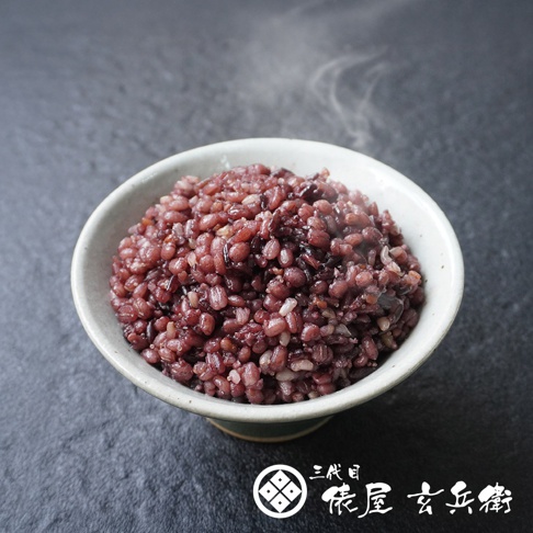 裝在淺色陶瓷碗中、冒著熱氣的深紫色十六穀米飯，顆粒飽滿並呈現豐富的紫紅色澤，碗底旁印有三代目俵屋玄兵衛的品牌標誌與文字，背景為深灰色的粗糙石紋質感，營造出溫暖且天然的氛圍。