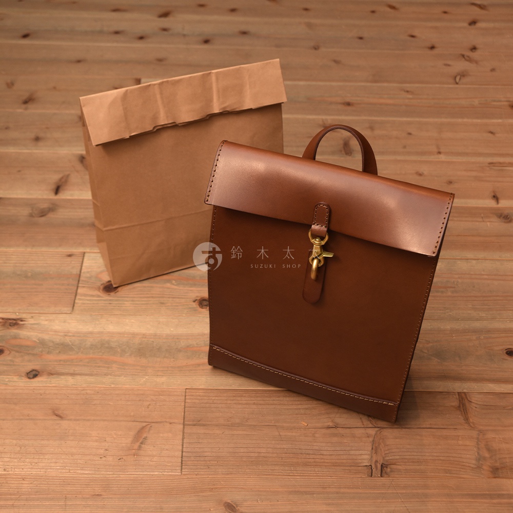 兩個職人手作皮革後背包，一個為棕色皮革，帶有金屬扣環；另一個為牛皮紙材質，呈長方形，兩者皆置於木質地板上。