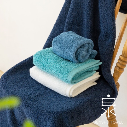 深藍色、藍綠色和白色浴巾疊放在椅子上，其中兩條浴巾捲起，展示了其柔軟的長纖維質地。