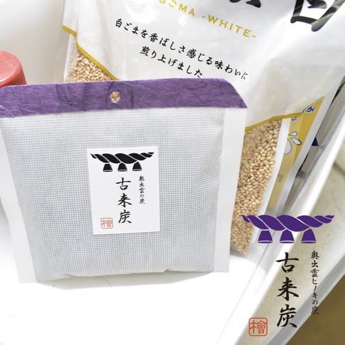 白色網狀包裝袋，上方有紫色紙質裝飾，中央貼有白色標籤，印有黑色日文「古來炭」字樣及圖騰。背景有裝有白色芝麻的透明包裝袋。