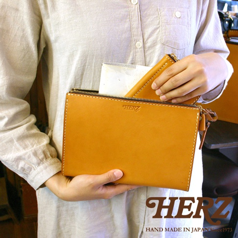 職人手作皮革三角底拉鍊手拿包，以天然皮革製成，呈現溫潤的焦糖色。簡潔的長方形設計，搭配同色系的車縫線和金屬拉鍊，展現細緻工藝。手拿包內有一小皮夾，方便收納。