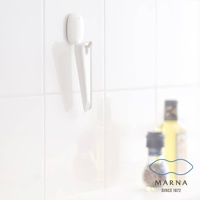 白色廚房清潔刮板，附白色掛座，掛於白色磁磚牆面上，刮板為塑膠材質，呈錐形，底部有斜切面。背景右側有兩瓶模糊的調味品。