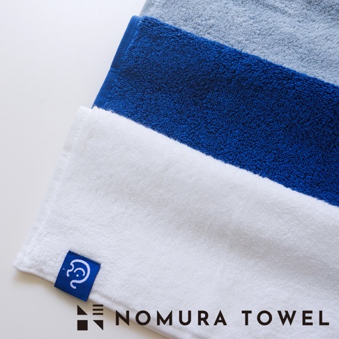 白色、藍色和灰色毛巾疊放，白色毛巾上印有NOMURA TOWEL的標誌