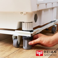 白色洗衣機下方放置著一個白色金屬可伸縮移動式平台，平台上裝有四個灰色滾輪和可調節高度的灰色底座，一隻手正在旋轉其中一個底座。