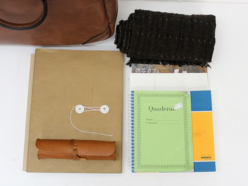 棕色皮革托特包，附有黑色皮革提把。旁邊擺放著深棕色紋理圍巾、牛皮紙文件夾、一個棕色皮革筆套，以及綠色封面筆記本和黃藍色封面筆記本。