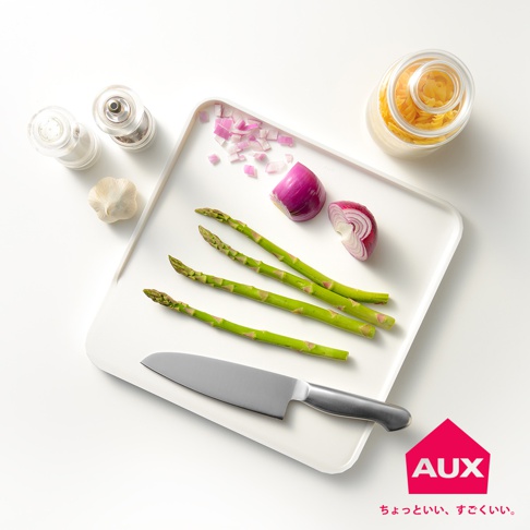 白色防溢大砧板上擺放著四根綠色蘆筍、切塊的紅洋蔥、半顆紅洋蔥、一顆大蒜、鹽罐、胡椒罐和一罐義大利麵。砧板左下角有一把銀色刀。