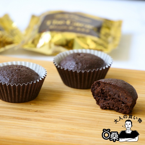 烤巧克力菓子禮盒(12入)：三個深棕色、表面略帶光澤的巧克力瑪芬，其中一個已咬開，露出濕潤的內部。瑪芬裝在深棕色帶有白色紙襯的紙杯中，擺放在淺木紋的背景上。後方有兩個金色包裝袋，模糊不清。