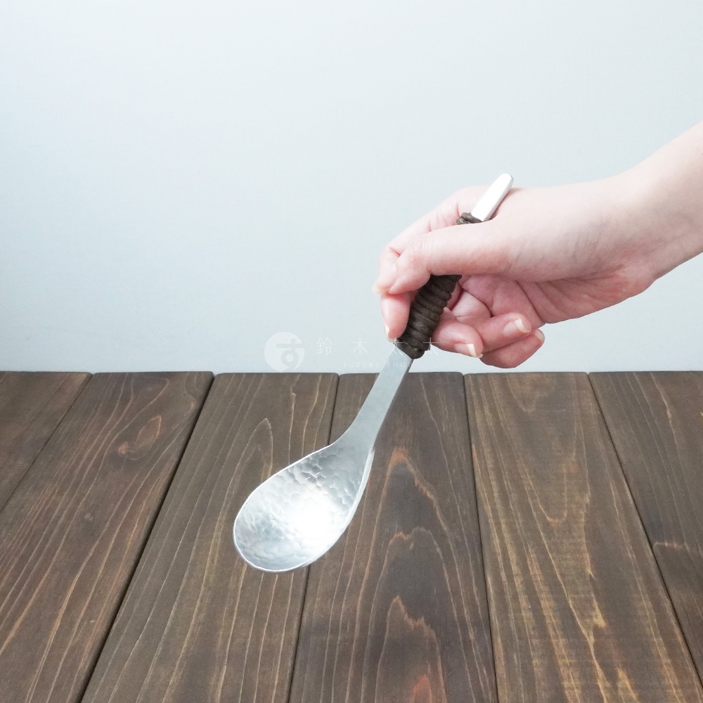 一手持鋁製湯勺，勺面帶有錘目紋，勺柄為深棕色纏繞設計。