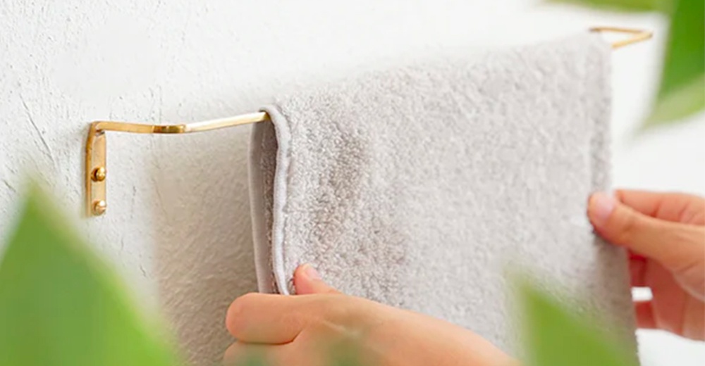 黃銅單桿毛巾架，黃銅材質，簡約設計，掛有淺灰色毛巾，毛巾架固定在白色牆面上。