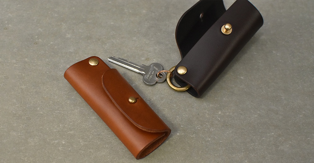 職人手作皮革鑰匙套，一個為棕褐色，另一個為深棕色，兩者皆配有金屬按扣及鑰匙圈。