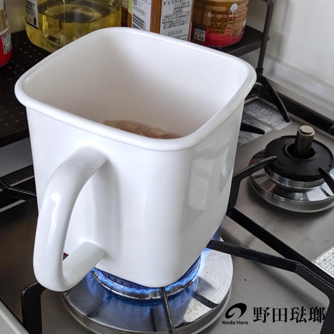 白色方形琺瑯儲物罐，帶有白色手柄，置於瓦斯爐上，底部有藍色火焰。