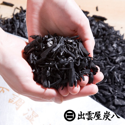 雙手捧著大量黑色、不規則形狀的木炭碎塊，背景是堆疊的木炭和印有文字的白色包裝。