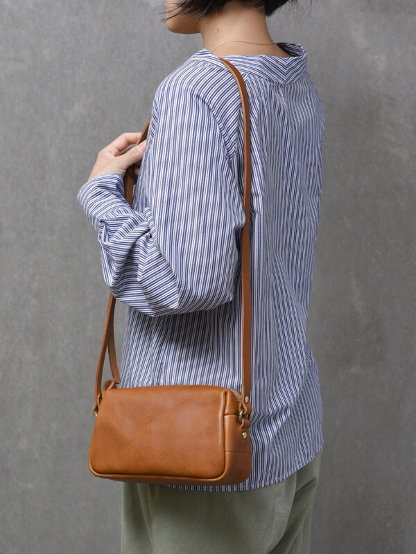 棕色皮革製成的輕巧拉鍊單肩包，包身呈現長方形，搭配一條細長的同色系皮革斜背帶，包包上有金屬拉鍊及裝飾釘。