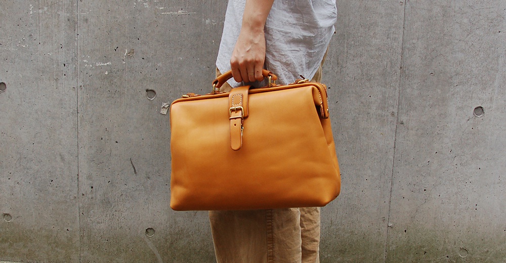 一只手提著一個駝色皮革軟式杜勒斯包，包包上有一個皮帶扣和金屬扣環，背景為水泥牆。