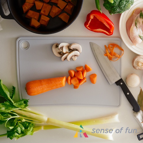 淺灰色矽膠砧板，上面放有切片蘑菇、胡蘿蔔塊、一根未切的胡蘿蔔以及一塊雞胸肉。旁邊還有一個刀具、紅椒、西蘭花和芹菜。