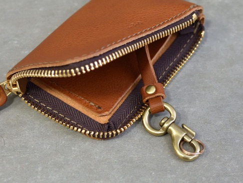 職人手作皮革Organ抽繩收納鑰匙包，採用溫暖的棕色皮革，搭配黃銅色拉鍊和掛鉤。包體為拉鍊開合設計，內層可見皮革內襯，外層有一個皮革抽繩連接金屬掛鉤。