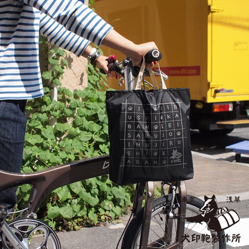 黑色環保折疊收納購物袋，印有白色日文文字與狗爪圖案，袋面設計為網格狀，兩側有米白色提把，裝在自行車前架上。