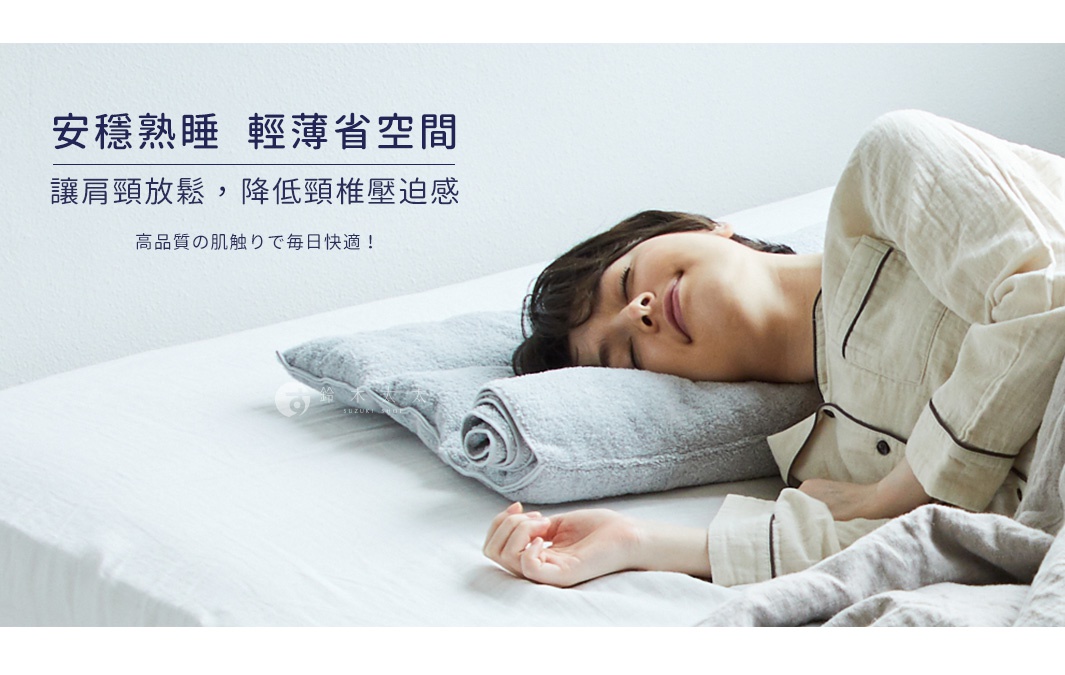 一個女子臉部特寫，閉著眼睛，微笑著，她枕在一個灰色的毛巾枕上，枕頭呈捲曲狀，枕頭上印有「鈴木雜貨」的字樣。