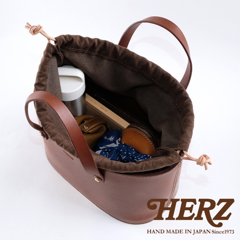 職人手作皮革束口手提包，深棕色皮革，內襯為深棕色帆布，束口處有繩結設計，包內裝有保溫瓶、筆記本、零錢包及藍白花紋布料。