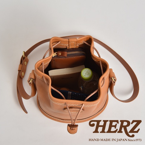 職人手作皮革束口單肩包，淺棕色，圓筒形設計，束口開合，內有錢包、書本、水瓶及藍白花紋布料。