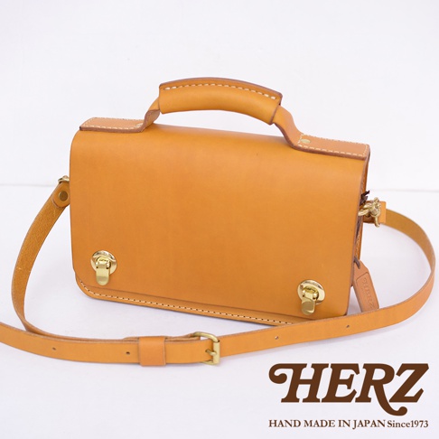 職人手作皮革經典兩用雙扣迷你單肩包，呈現溫潤的駝色，包身結構方正，頂部設有提把，附有可調節的長斜背帶，正面有雙金屬鎖扣設計，材質為天然皮革。
