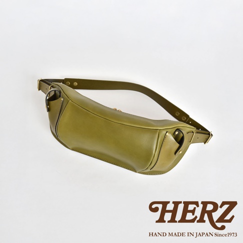 HERZ 品牌標誌，下方有 HAND MADE IN JAPAN Since1973 字樣，圖片展示一個綠色皮革製成的斜背包，背包呈現彎月形，底部邊緣有品牌標誌。