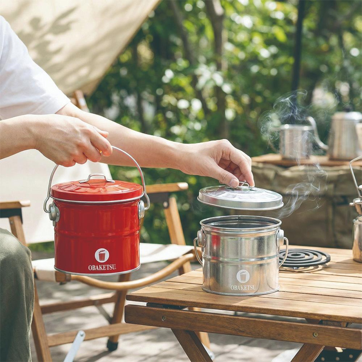 OBAKETSU 手工製蚊香桶，紅色款與銀色款並列於木桌上，兩款皆有提把和蓋子，銀色款上冒出陣陣煙霧，展現其功能性。