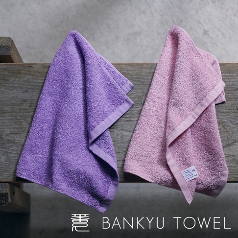 兩條「匠心獨技瞬吸乾爽毛巾」，一條為紫色，另一條為粉紅色，均為柔軟材質，邊緣有同色系滾邊，懸掛在木質架上。