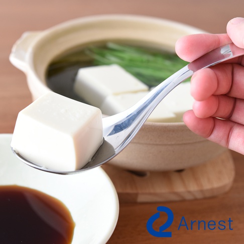 手持一只不鏽鋼瀝水湯匙，湯匙舀著一塊白色豆腐，湯匙上可見細小的孔洞。湯匙後方是裝有豆腐、綠色蔥段和湯汁的陶鍋，鍋子下方有木製隔熱墊。