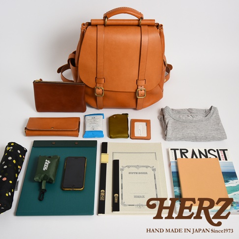 日本職人品牌 HERZ 棕色皮革後背包平鋪展示，上方為屋瓦風雙扣設計的皮背包，下方陳列日常收納物，包括錢包、筆記本、手機、折疊傘、衣服及雜誌等隨身用品，整體呈現日系簡約質感與手作皮革風格。