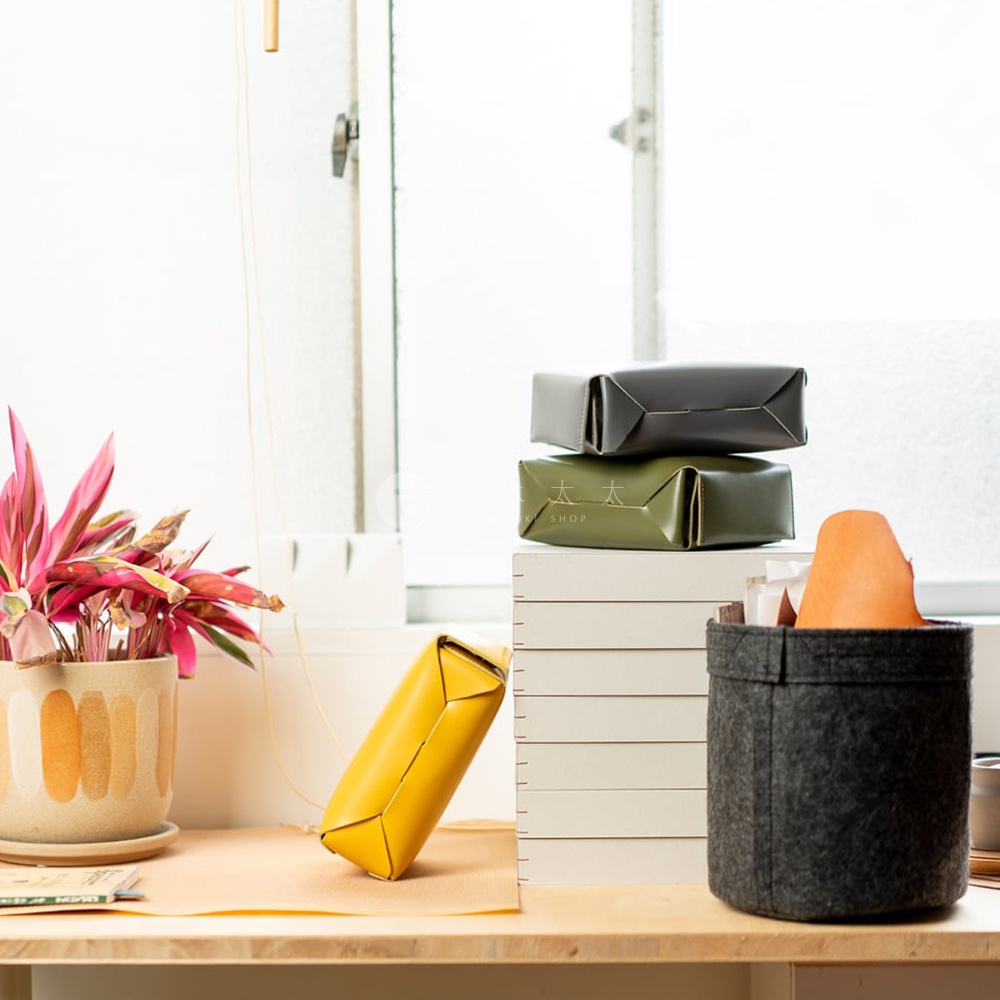 綠色和灰色皮包堆疊在白色盒子上，旁邊有一個灰色毛氈收納籃，籃子裡裝著橘色的皮件。