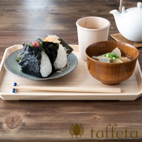 淺色楓木托盤上擺放著一盤飯糰、一碗味噌湯和一個木杯。飯糰裹著海苔，裝在灰綠色盤子裡。味噌湯盛在木碗中，碗裡有白蘿蔔和蔥花。木杯裡裝著茶。